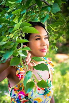 A beautiful mixed race African American woman in yellow floral dress standing in the forest on a sunny summer day parts a tree branch covered in leaves with her hand.