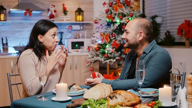 Shocked woman getting asked to marry partner proposing with engagement ring on christmas eve. Couple engaged and enjoying festive dinner. People celebrating holiday and proposal