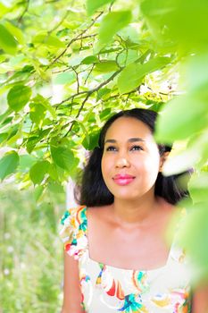 Beautiful bright portrait of a mixed race African American woman standing under a tree with bright green leaves on a sunny summer day as she wears a yellow floral dress and looks up and to the side.