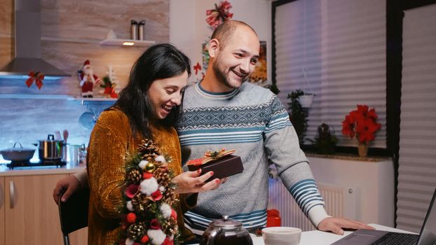 Man and woman receiving gift on video call communication for christmas eve celebration. Festive couple with present box celebrating winter holiday with family on online conference