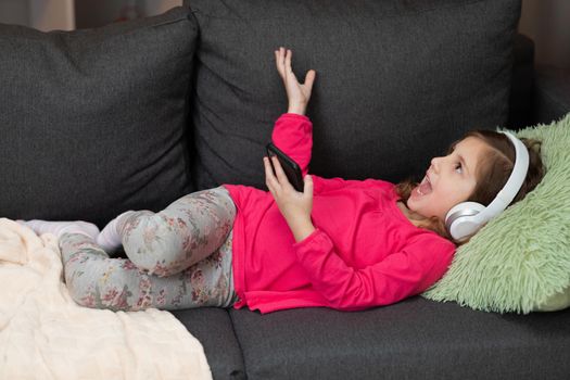 Happy Little girl in wireless headphones chilling laughing on sofa listening to favorite music holding phone using mobile online player app. Enjoy peaceful mood wearing earphones at home