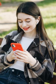 Young beautiful girl listens to music and leaves through the tape of social networks on her smartphone while sitting on a bench in the park.