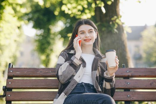 Attractive young woman dressed in stylish wear having phone conversation with boyfriend. Discussing ideas for weekends during work break sitting outdoors on city bench with Coffee.