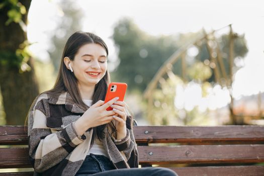 Happy girl listening to music in the park using mobile phone and wearing headphones. Attractive woman browsing on mobile phone in public park. Cute young girl in urban background listening to music