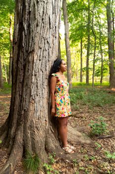 Full body portrait of a beautiful happy mixed race African American woman wearing a yellow floral dress standing and leaning against a large tree trunk in a forest and stares off into the distance.