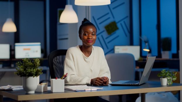 Portrait of business black woman looking at camera smiling raising head from laptop working in start-up financial company late at night. Focused employee doing overtime respecting deadline of project