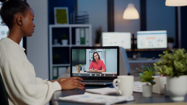 African businesswoman discussing with remotely woman partner online sitting in front of laptop working in start-up office talking on video call during virtual meeting at midnight, using headphone