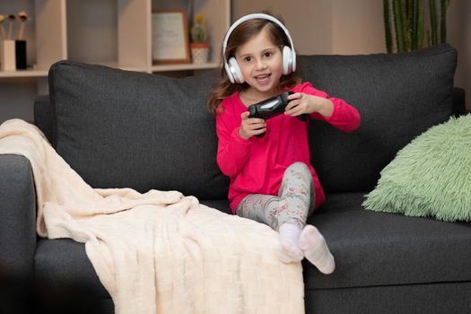 Young Little girl sitting on sofa playing a video in living room at home. Excited gamer girl hand holding joystick playing console game using a wireless controller. Having fun,enjoy happy expression.