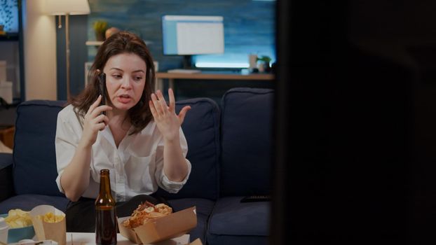 Annoyed woman talking on smartphone with boss after work at home while trying to eat takeaway food from delivery boxes. Person with fast food meal on table using phone for work call