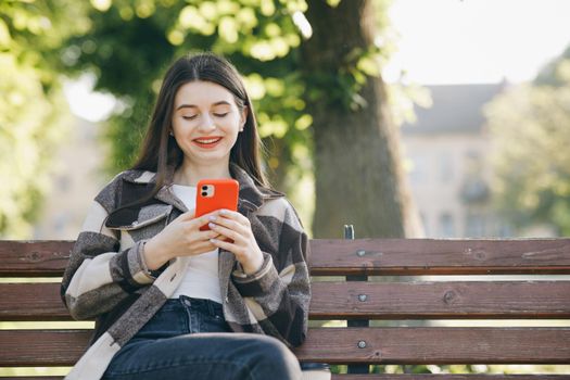 Pretty young woman in trendy casual outfit sits on a bench in the city park, actively texts messages via her phone. Being online, modern technologies.