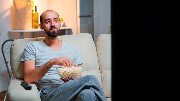 Man late at night in his apartment looking at TV entertainment, laughing and having fun while eating popcorn snack. Smiling caucasian person leisure time on the sofa in front of television