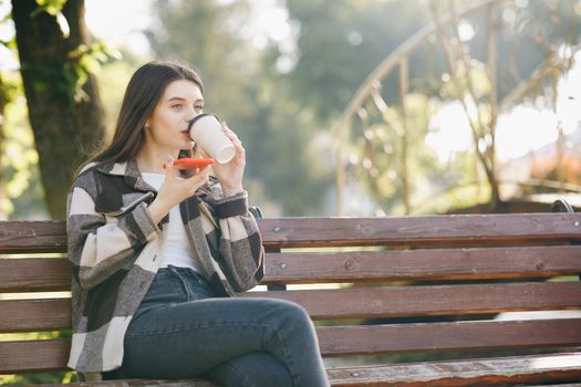 Pretty young woman in trendy casual outfit sits on a bench in the city park, actively texts messages via her phone. Being online, modern technologies. Holding paper Cup with hot Drink in her Hands.