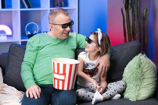 Elderly man with a little girl Wearing 3d Glasses watching tv andeating popcorn. Senior, old generation, grandphather family time relax with young girl kid on sofa in living room concept.