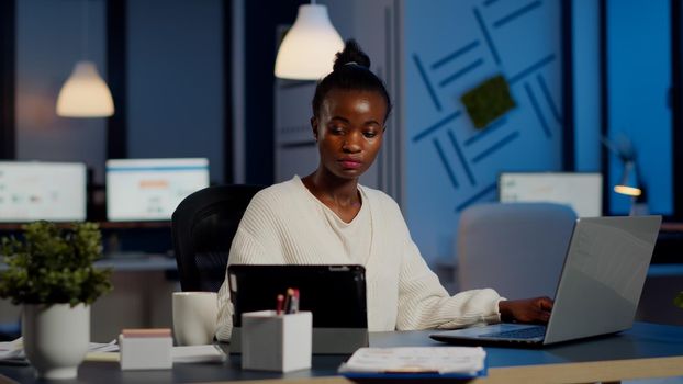Multitasking black business woman working at laptop and tablet in same time doing overtime in start-up office. Busy african employee analysing financial statistics overworking writing, searching.