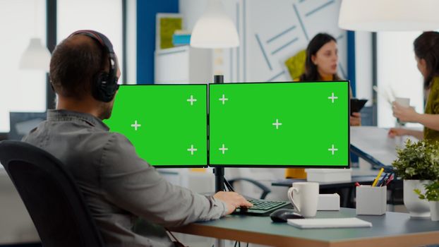 Employee with headphones using dual monitror setup with green screen, chroma key mock up isolated display sitting in video production studio. Man ditor processing film montage on pc in creative agency