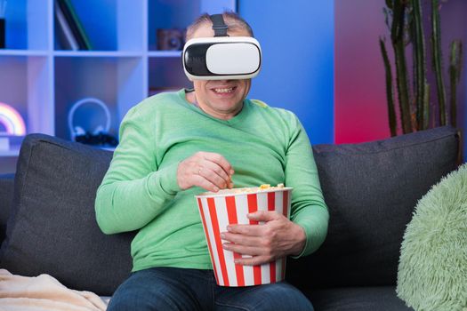 Senior man sitting on Sofa and holding popcorn, He wearing virtual reality glasses and lift up his hand to selective with VR Screen