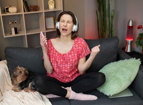 Joyful girl in headphones is dance on sofa and using smartphone touching screen. Modern technology concept.