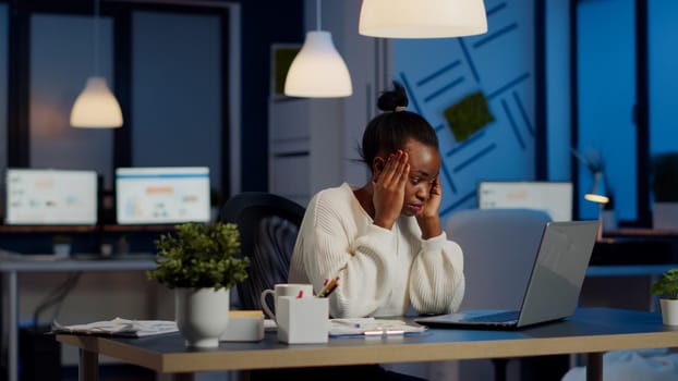 Overworked african manager woman massaging head while sitting at workplace in start-up office working overtime at laptop. Exhausted tensed businesswoman respecting deadline coping with migraine