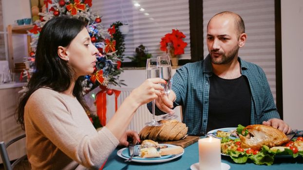 Man and woman clinking glasses of champagne at festive dinner, celebrating christmas eve with chicken and alcohol. Couple enjoying meal for traditional holiday celebration. People eating