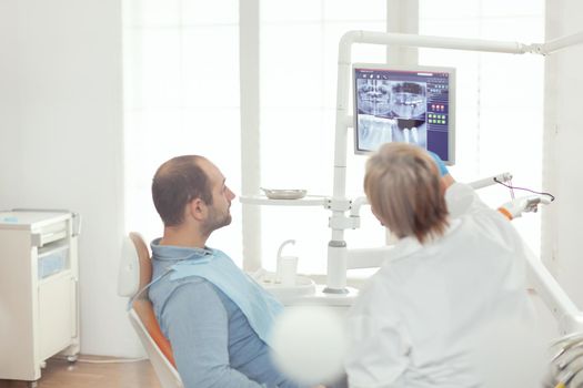 Sick man sitting on dental chair looking at teeth radiography preparing for somatology surgery. Senior doctor explaining toothache pain preparing medical treatment while working in orthodontic office