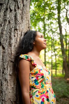 Beautiful portrait of a happy mixed race African American woman wearing a yellow floral dress leaning against a large rough bark covered tree trunk in a forest and looks up into the distance.