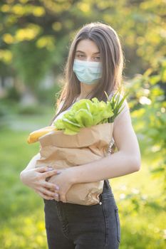 Vertical Portrait of a courier in a medical mask holding a parcel. Caucasian young pretty woman delivery worker in medical mask on the park carrying packet with fresh food.
