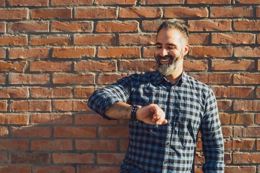 Happy modern businessman with beard looking at his watch while standing in front of brick wall outdoor.