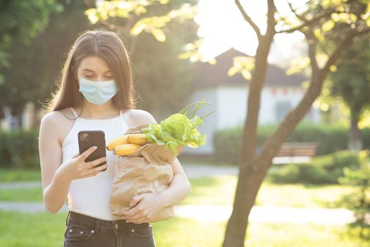 Caucasian young pretty woman in medical mask walking the street carrying packet with fresh food. Female beautiful going to deliver grocery.