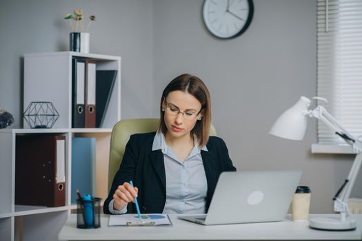 Businesswoman Working on a Laptop in Her Modern Office Holding Paper Financial Report. Confident Businesswoman Working With Data From Financial Documents and Typing on Computer in Office.