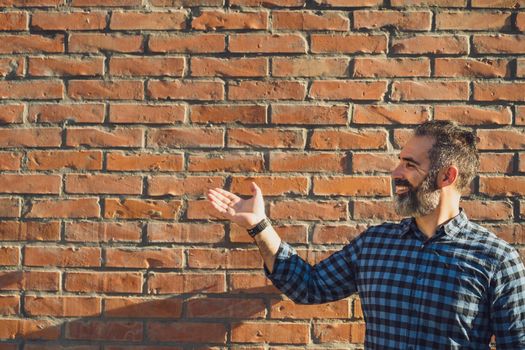 Portrait of modern businessman with beard gesturing while standing in front of brick wall outdoor.