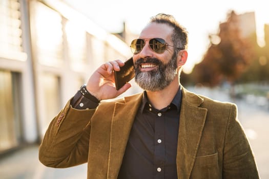 Modern businessman with beard using mobile phone while standing on the city street.