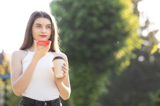 Young girl walking in the Park speaking on Smartphone. Happy girl using a smart phone voice recognition audio ai message speech function on line walking in park. Having cup with Coffee in her Hands