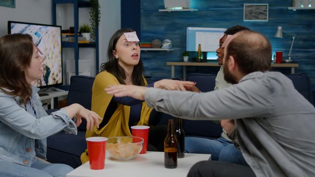 Mixed race friends playing Guess who game with sticky papers attached on forehead. Group of multiracial people having fun, laughing together while sitting on sofa in living room late at night