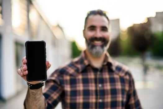 Modern businessman with beard showing blank screen of phone while standing on the city street. Focus on the phone.