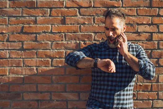 Businessman in panic with beard looking at watch while standing in front of brick wall outdoor.