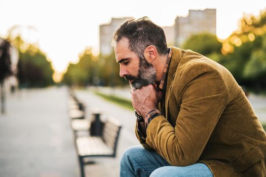 Depressed businessman with beard sitting on the bench in the city.