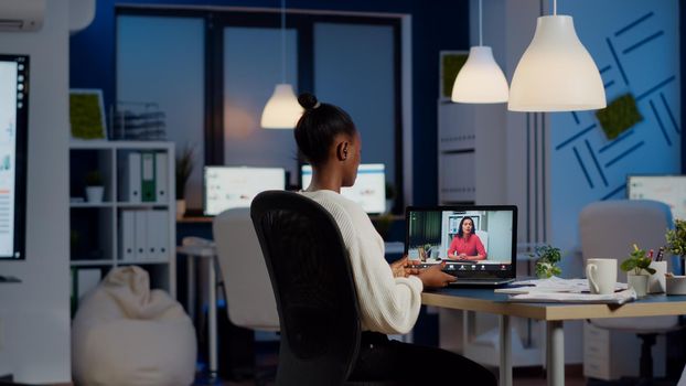 African freelancer working remotely discussing with woman partner online sitting in front of laptop in start-up office talking during virtual meeting at midnight, using headphone doing overtime