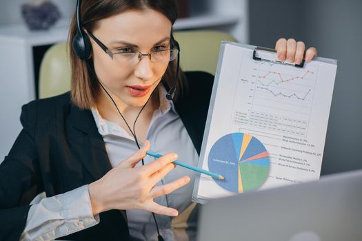 Businesswoman in Headphones Holding Paper Financial Report talk at Webcam make video call in office, business coach looking at camera speak show statistics explain marketing strategy for client.