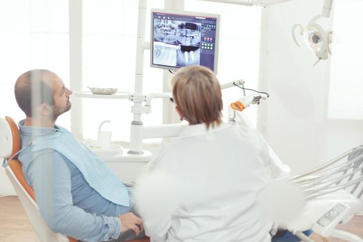 Senior stomatologist woman discussing with sick man about healdcare treatment looking at tooth treatment during stomatology hospital office. Orthodontic doctor examining dental x-ray