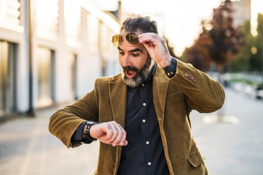 Businessman with beard in panic looking at watch and walking in hurry on the city street.