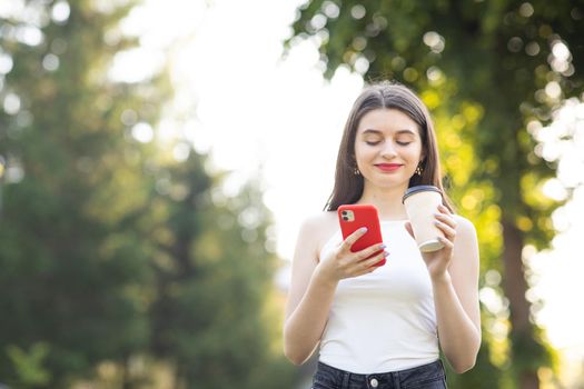 Girl Laughing while looking on her Smartphone. Town Landscape with trees. Cheerful young girl with a paper Cup of coffee in her left hand.