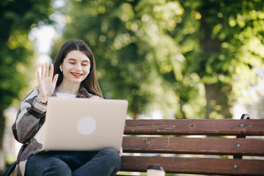 Girl is waving her hand to webcam on laptop while sitting on park bench. Student using webcam on laptop has video chat with friend. Caucasian woman smiling sitting on the bench lunch time outdoors