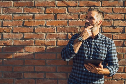 Portrait of modern businessman with beard thinking and using digital tablet while standing in front of brick wall outdoor.