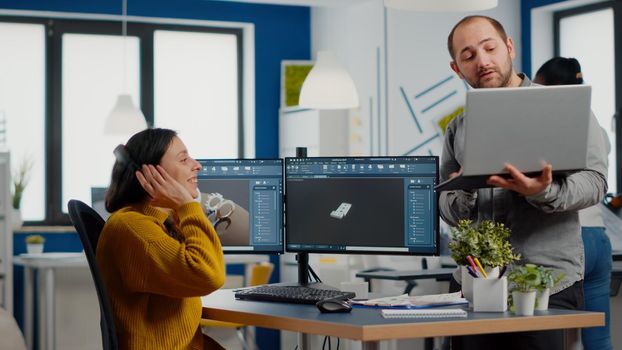 Industrial designer discussing with woman engineer while working in CAD program, designing new components, comparing 3D Prototype with project from laptop. Workteam using computer with two monitors