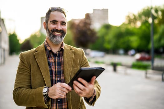Portrait of modern businessman with beard using digital tablet while standing in on the city street.