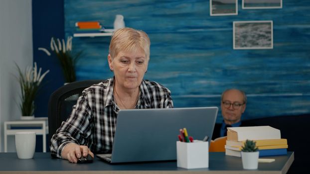 Mature caucasian lady working on laptop at home workplace, typing on computer drinking coffee. Experienced manager doing financial project during self isolation while husband reading a book in background