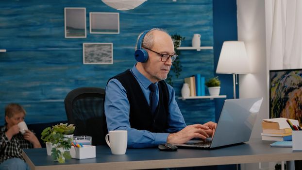 Focused old entrepreneur with headphones listening music while checking graphics writing on laptop working from home. Retired man using modern technology reading typing, searching on computer