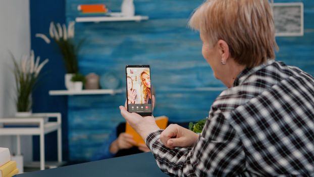 Senior woman having video call online talk with nephew using smartphone sitting in living room. Retired granny using modern internet online tech to connect with grandparents