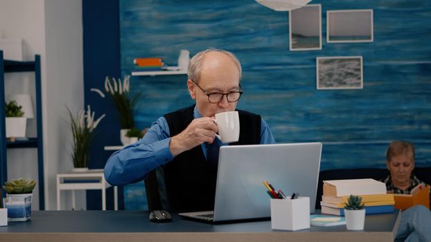 Senior businessman reading reports sitting in front of laptop working from home drinking coffee. Retired man using modern technology analysing typing searching while wife sitting on couch reading a book