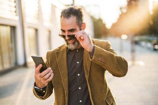 Modern businessman with beard using mobile phone while walking on the city street
